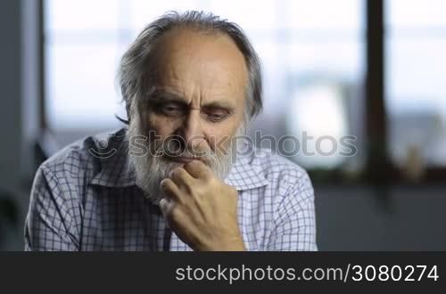 Portrait of pensive handsome senior man stroking his beard and pondering over life. Thoughtful old man thinking about days go by, remembering best moments and ironicaly smiling over wide window background.