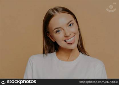 Portrait of playful dark haired woman tilts head shows tongue and smiles positively has fun indoor dressed in white sweater poses against beige background. Human face expressions and positive feelings