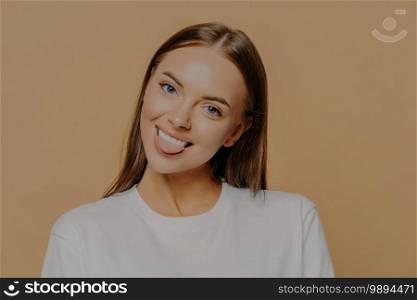 Portrait of playful dark haired woman tilts head shows tongue and smiles positively has fun indoor dressed in white sweater poses against beige background. Human face expressions and positive feelings