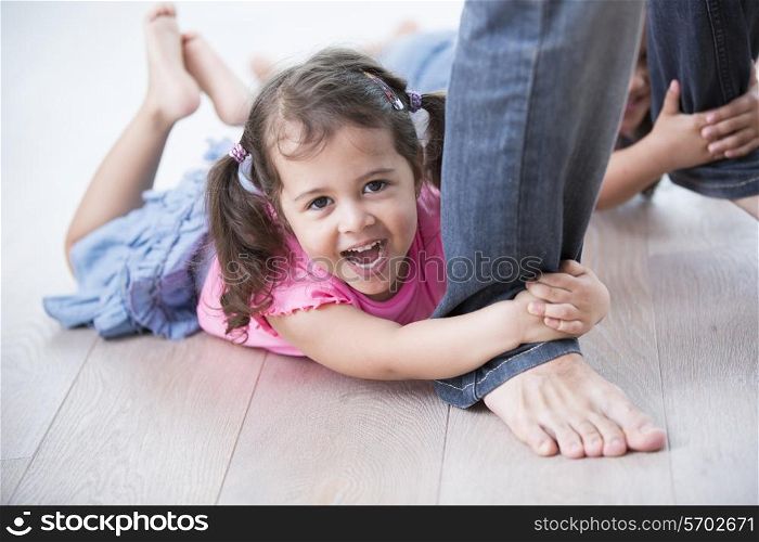 Portrait of playful girl with sister holding father&rsquo;s legs on hardwood floor