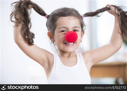 Portrait of playful little girl wearing clown nose holding pigtails at home
