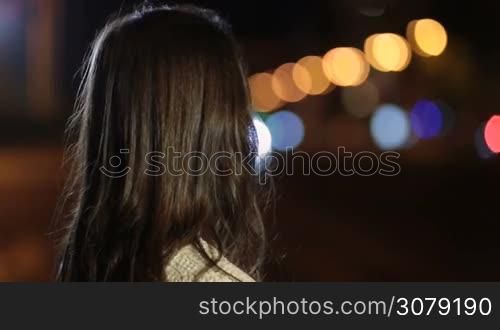 Portrait of playful sexy brunette woman flirting with someone, turning her head back and looking with seductive toothy smile against colorful bokeh of night city streetlights. Charming girl posing and smiling on street at night.