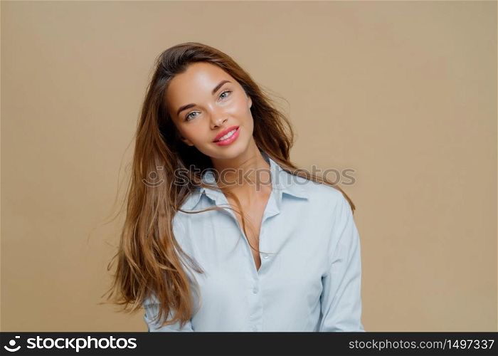 Portrait of pleasant looking cheerful female has long hair, tilts head and looks with smile at camera, wears blue shirt, uses cosmetics for makeup, poses against brown background, blank space aside