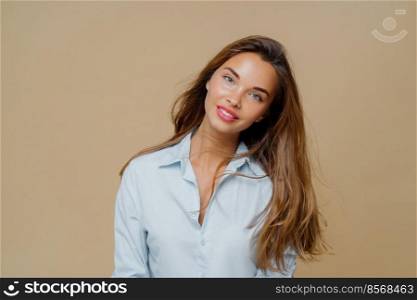 Portrait of pleasant looking cheerful female has long hair, tilts head and looks with smile at camera, wears blue shirt, uses cosmetics for makeup, poses against brown background, blank space aside