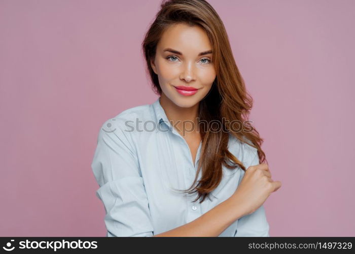 Portrait of pleasant looking female model with dark long hair, looks confidently at camera, has makeup, smiles gently, poses to make photo for journal cover, isolated over purple background.