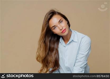 Portrait of pleasant looking female tilts head on right, has long well cared hair, wears blue shirt, has appealing appearance, makeup, isolated over brown background with blank space for information