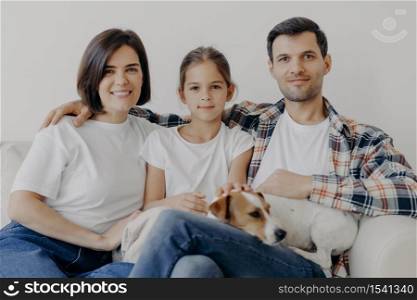 Portrait of pleasant looking mother, dad and small child, sleeping pet, pose all together at sofa against white wall, dressed in casual clothes, spend weekend at home, have friendly relationship