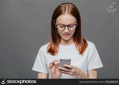 Portrait of pleasant looking relaxed woman holds modern cell phone, types text message on cell phone, connected to wirelesss internet, dressed in white t shirt, isolated over grey wall. Technology
