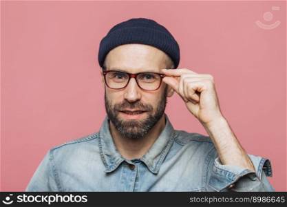 Portrait of pleasant looking satisfied bearded man with pleasant appearance looks confidently through spectacles, wears fashionable clothing, poses against pink studio background. Facial expressions