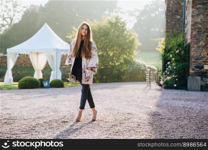 Portrait of pleasant looking slim tall woman wears cape and high heeled shoes, stands crossed legs, poses outdoors against ancient area, has confident look directly into camera. Lifestyle concept