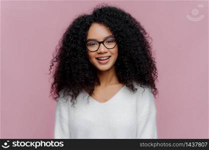 Portrait of pleasant looking smiling woman with Afro hairstyle, wears optical glasses and white sweater, has cheerful face expression, isolated over purple background. Millennial girl poses indoor
