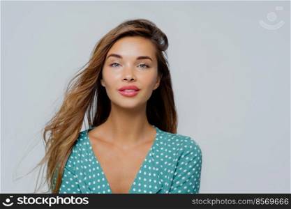 Portrait of pleasant looking young woman looks calmly at camera, has long hair, dressed in polka dot blouse, poses against white background, has natural beauty, has well cared face and makeup