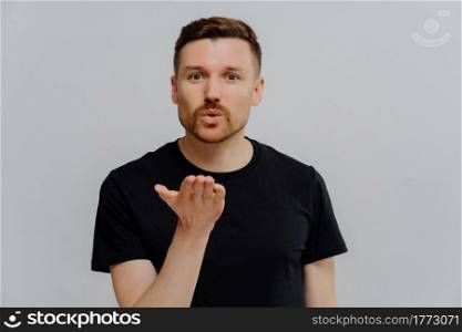 Portrait of pleasant young man in basic black t shirt sending air kiss and looking at camera, demonstrating feelings while standing isolated over grey studio background. Love expression concept. Young guy blowing air kiss to his girlfriend and demonstrating feelings