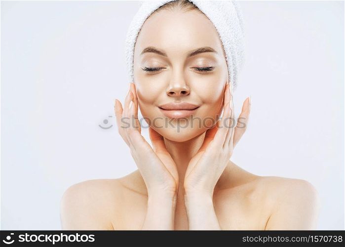 Portrait of pleased European woman closes eyes, stands half naked with wrapped towel on head, has healthy soft skin, poses bare shoulders, stands indoor. Women beauty, personal care concept.