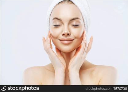 Portrait of pleased European woman closes eyes, stands half naked with wrapped towel on head, has healthy soft skin, poses bare shoulders, stands indoor. Women beauty, personal care concept.