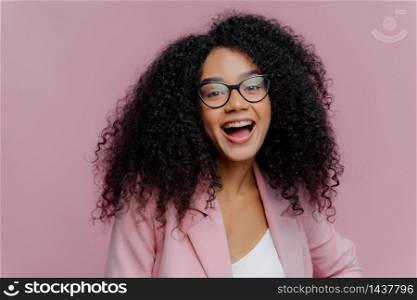Portrait of pleased young curly haired woman feels overemotive, keeps mouth widely opened, wears optical glasses and formal suit, laughs happily at news, poses indoor against bright background
