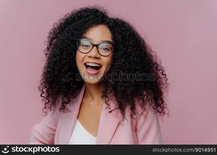 Portrait of pleased young curly haired woman feels overemotive, keeps mouth widely opened, wears optical glasses and formal suit, laughs happily at news, poses indoor against bright background
