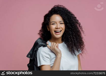 Portrait of positive African American woman smiles toothily, being in good mood after walk in park, dressed in white t shirt holds leather jacket on shoulder isolated on pink background. People, style