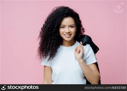 Portrait of positive ethnic woman wears white t shirt, carries jacket on shoulder, has cheerful expression, ready for picnic, enjoys day off, poses indoor against rosy background. People, lifestyle