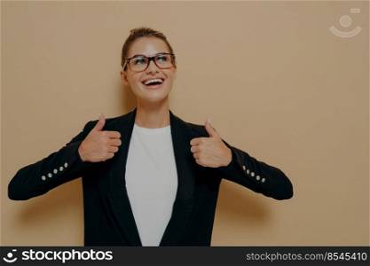 Portrait of positive female customer in eyeglasses with broad smile showing thumbs up with both hands and looking at copy space with happy expression while posing isolated over beige background. Positive female customer in eyeglasses with broad smile showing thumbs up with both hands
