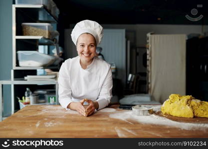 Portrait of positive smiling and satisfied woman baker in uniform leaned on wooden table with dough. Bakery house female worker. Portrait of positive baker in uniform leaned on wooden table with dough