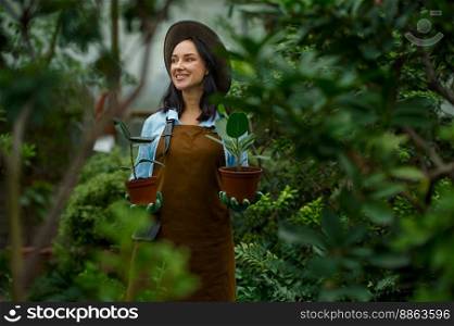 Portrait of positive smiling woman gardener wearing apron carrying flowerpots in hand selective focus with view through lush greenhouse foliage. Positive smiling woman gardener carrying flowerpots in hand selective focus