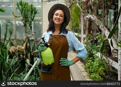 Portrait of positive smiling young woman gardener with pulverizer sprayer in hand posing for camera over greenhouse garden background. Portrait of young woman gardener with pulverizer sprayer in hand