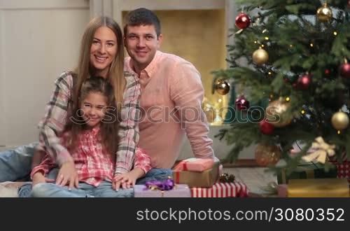 Portrait of positive young family with lovely daughter waving hands and smiling over Christmas decorated room background. Attractive parents and little girl sitting together near xmas tree during winter holidays, looking at camera and waving hands.