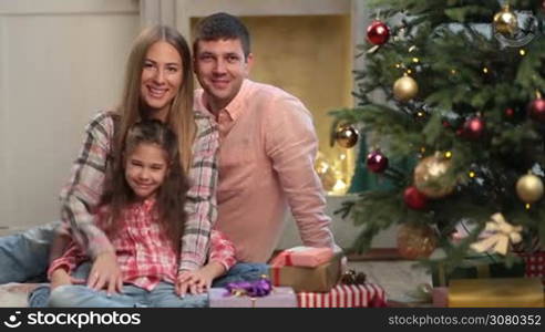 Portrait of positive young family with lovely daughter waving hands and smiling over Christmas decorated room background. Attractive parents and little girl sitting together near xmas tree during winter holidays, looking at camera and waving hands.