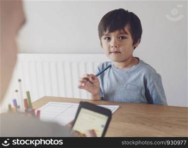 Portrait of preschool kid holding pencil with blurry foreground of a man typing on mobile phone, Selective focus of Child doing homework with blurry foreground of father searching internet on smart phone.