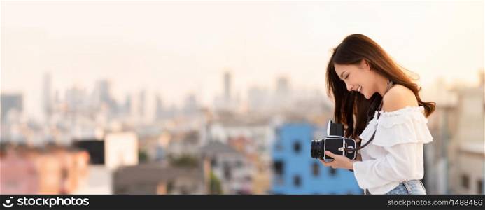 Portrait of Pretty Asian woman standing at outside while holding retro camera with Buildings background. Happy, Having Fun. Banner, Panoramic.