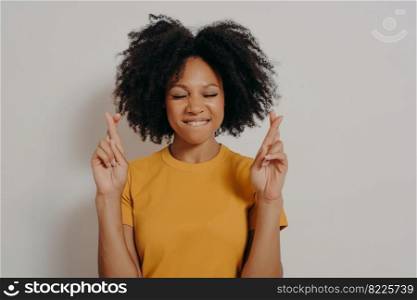 Portrait of pretty cheerful african girl standing isolated over white studio background with copy space, holding fingers crossed for good luck, with closed eyes biting lower lip while making wish. Portrait of pretty cheerful african girl standing in studio and holding fingers crossed for luck