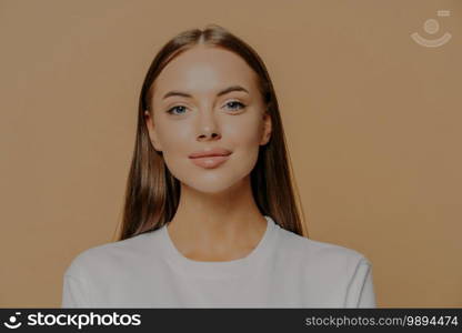 Portrait of pretty European woman with long hair looks confidently ar camera has healthy glowing skin makeup wears casual jumper isolated over brown studio wall. Beauty and wellness concept.