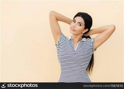 Portrait of pretty female with dark hair, makes pony tail, dressed in casual striped t shirt, looks seriously at camera, poses against studio wall with blank space for your advetisement or promotion