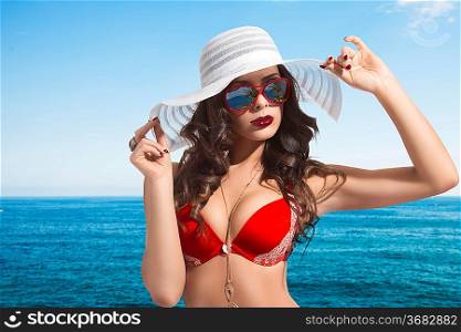 portrait of pretty girl with brown wavy hair, red sexy bikini, white summer hat and sunglasses near the sea