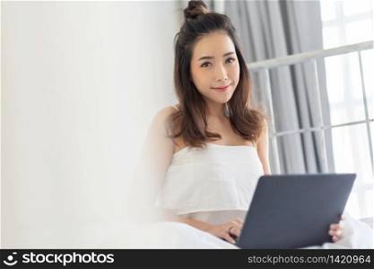 Portrait of pretty woman sitting in bed and smiling and looking at camera on morning. Positive Asian beautiful girl using laptop computer in white bedroom at home on holiday.