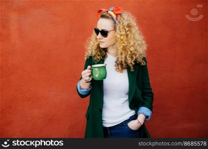 Portrait of pretty woman with curly hair wearing headband, stylish sunglasses and green jacket holding green cup of tea in one hand and other hand in pocket looking aside isolated over orange wall