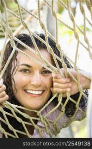 Portrait of pretty young adult Caucasian brunette female peeking through hammock weave and smiling at viewer.