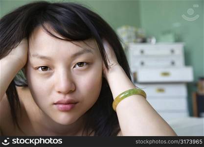 Portrait of pretty young Asian woman lying in bed with hands in hair making eye contact.