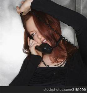 Portrait of pretty young redhead woman holding telephone receiver to ear with cord wrapped around head smiling.