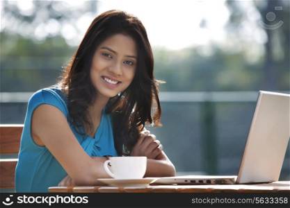 Portrait of pretty young woman smiling while sitting at table with laptop and cup of coffee