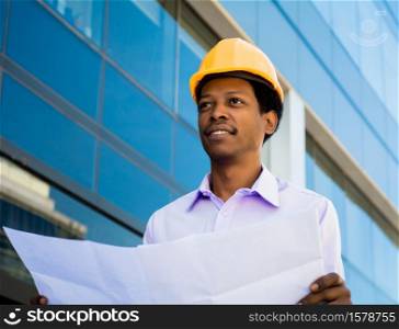 Portrait of professional architect in helmet looking at blue prints outside modern building. Engineer and architect concept.