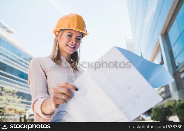Portrait of professional architect wearing yellow helmet and looking at blue prints outside modern building. Engineer and architect concept.
