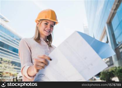 Portrait of professional architect wearing yellow helmet and looking at blue prints outside modern building. Engineer and architect concept.