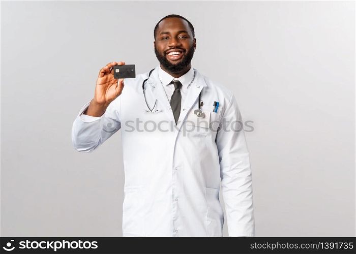 Portrait of professional, handsome african-american doctor recommend paying for medical insurance, use bank service, showing credit card with satisfied, happy face, grey background.. Portrait of professional, handsome african-american doctor recommend paying for medical insurance, use bank service, showing credit card with satisfied, happy face, grey background