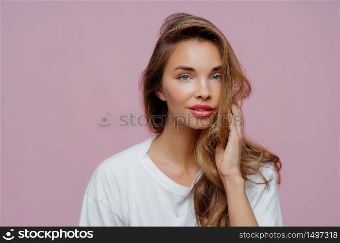 Portrait of relaxed beautiful Caucasian lady has blue eyes, well cared skin, looks directly at camera, has long hair, minimal makeup, wears casual white comfortable jumper, models in studio.