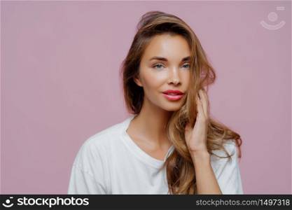 Portrait of relaxed beautiful Caucasian lady has blue eyes, well cared skin, looks directly at camera, has long hair, minimal makeup, wears casual white comfortable jumper, models in studio.