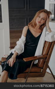 Portrait of relaxed caucasian woman posing looking at camera sitting on cozy chair indoors, stylish pretty young female fashion model wearing black dress, spends leisure alone, relaxing at home.. Stylish pretty young caucasian female wearing black dress sitting on cozy chair relaxing at home