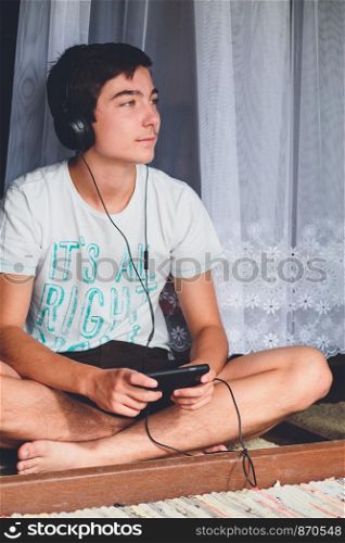Portrait of relaxed young boy sitting at the patio door and listening to music from mobile phone