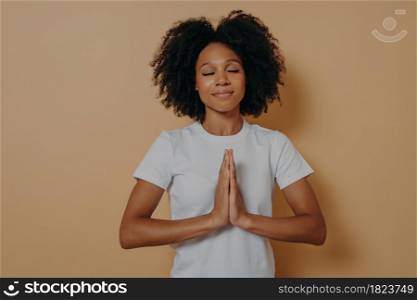 Portrait of religious faithful african american young woman holding hands clasped together and praying while standing isolated over brown background, hopeful female with eyes closed asking about help. African american young woman holding hands clasped together and praying on beige background